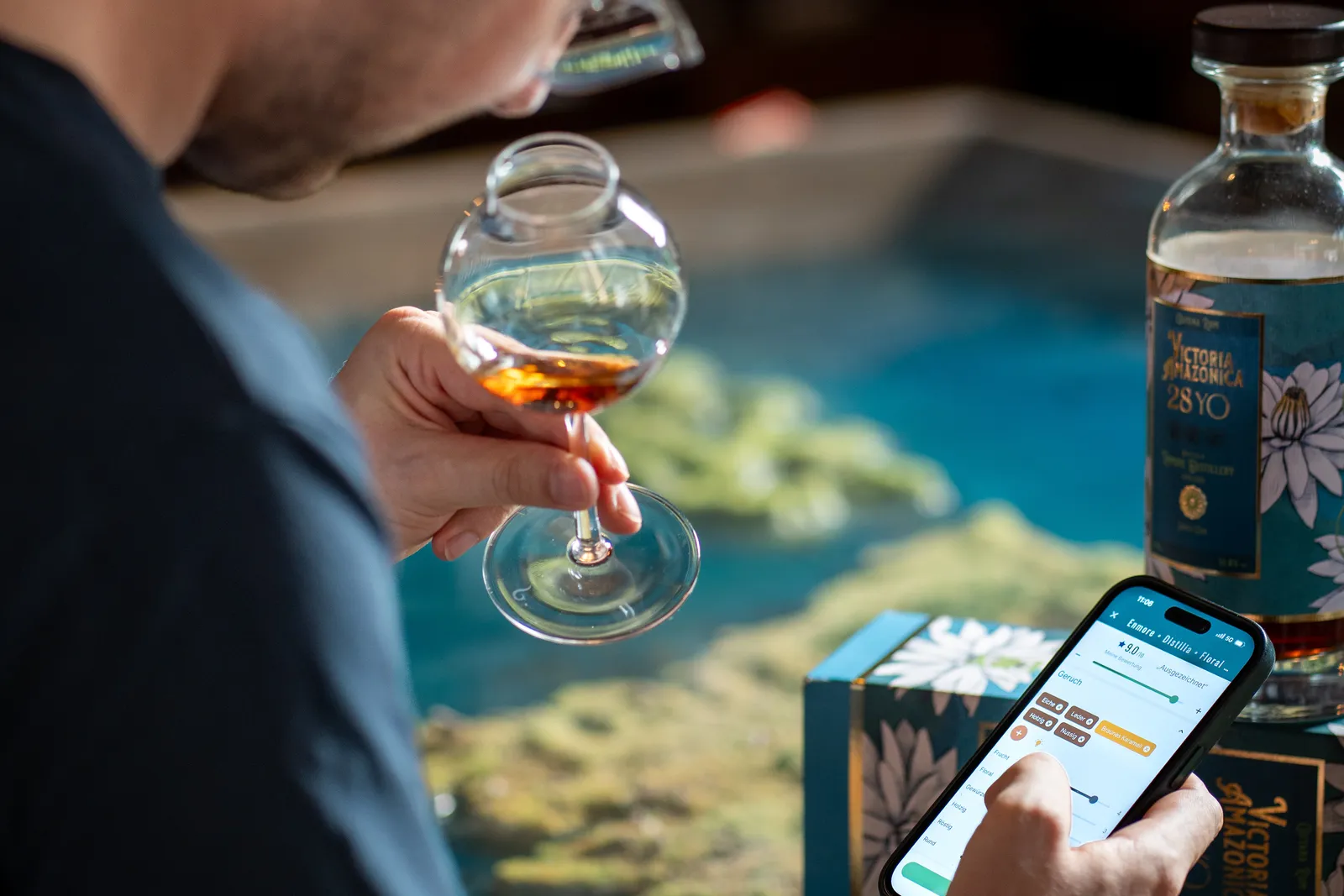 A RumX user nosing an amber rum from a tasting glass while logging it in the RumX app, a 28-year-old rum bottle beside them