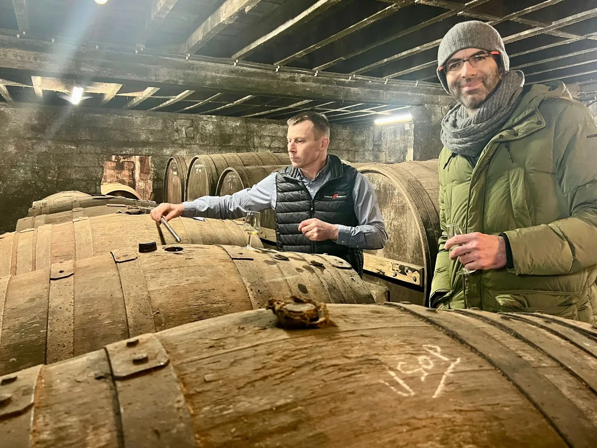 Two men on top of stacked casks in a dark warehouse, one drawing a sample, with a shaft of daylight behind them.
