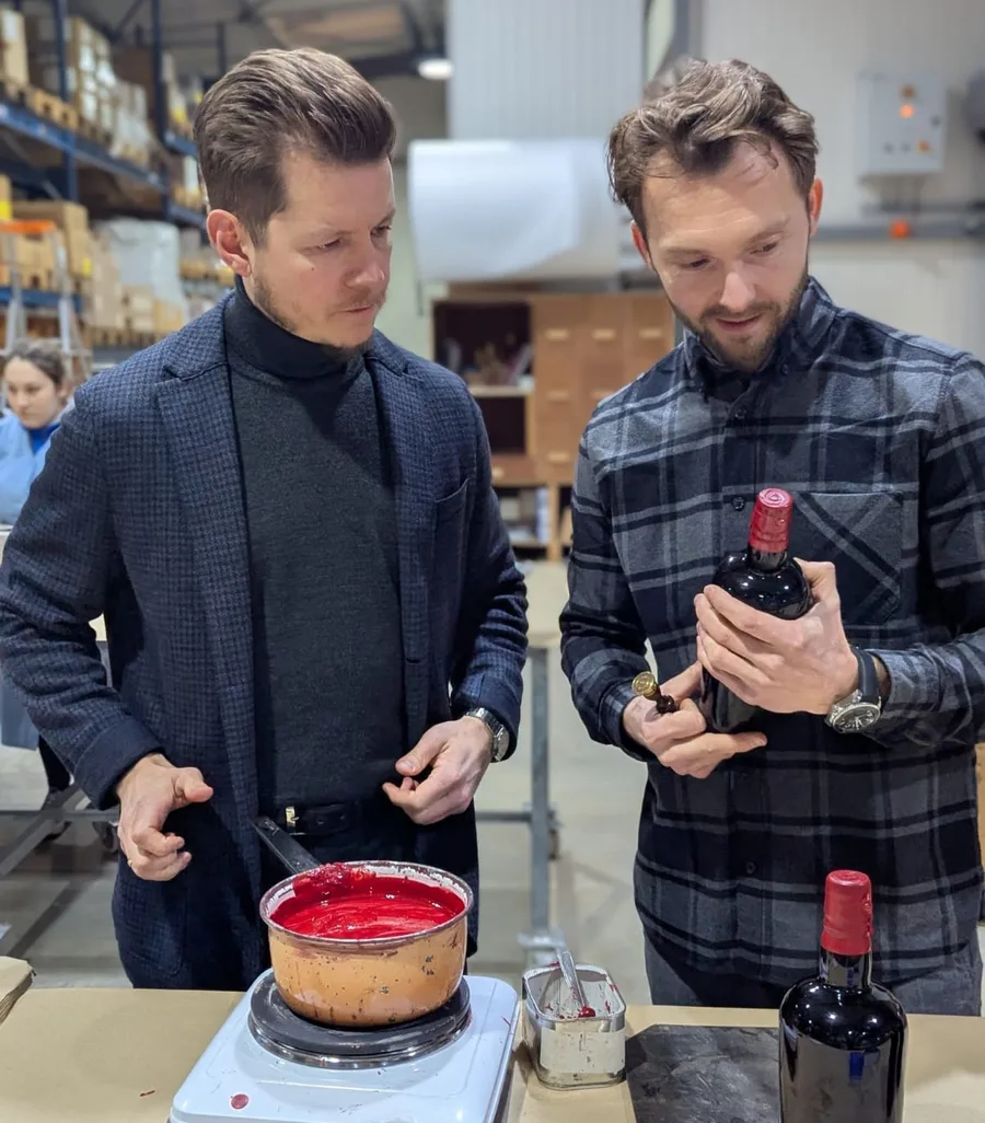 Two men in a bottling hall inspecting a bottle next to a pot of red sealing wax on a scale.