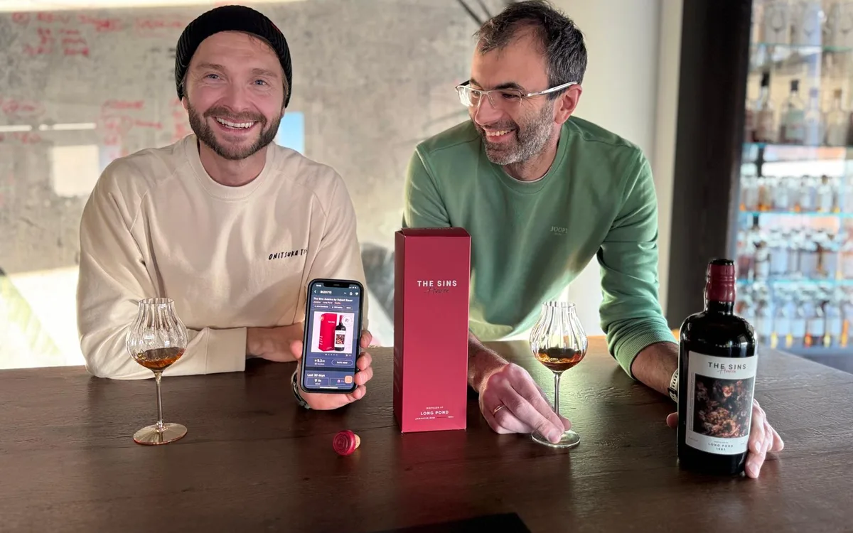 The two Distilia founders at a tasting table with the RumX app open on a phone, next to a bottle and box of The Sins Avarice.