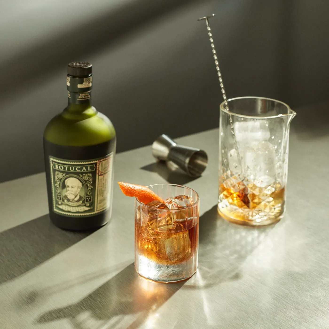 Bottle of Diplomático Reserva Exclusiva with its Botucal label next to an Old Fashioned with orange zest and a mixing glass with ice