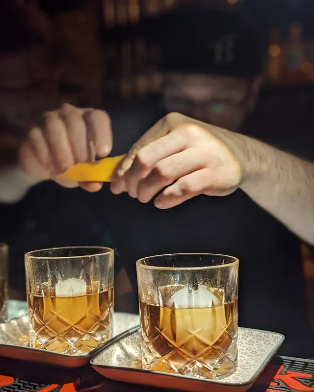 Bartender squeezing a citrus zest over two Old Fashioneds with large ice cubes