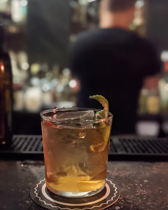 Old Fashioned with orange zest on a dark bar counter, bartender blurred in the background