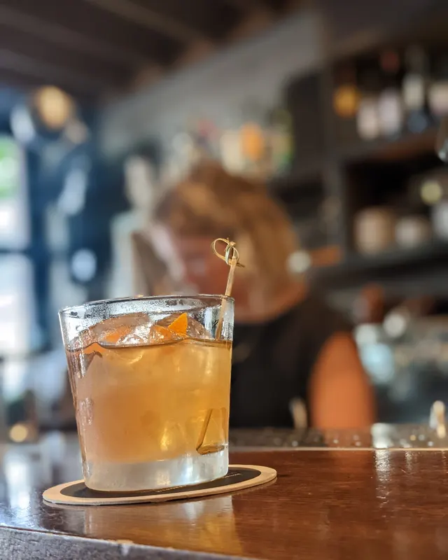 Old Fashioned with an orange garnish on a pick in front of a lit back bar