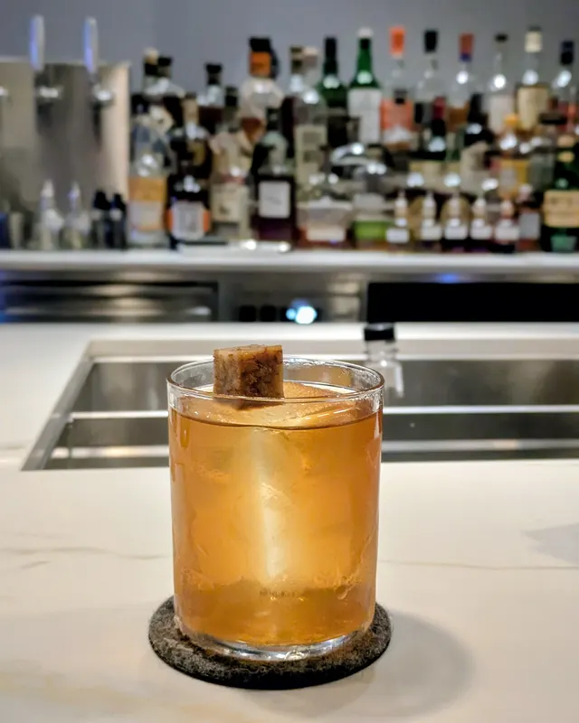 Old Fashioned with a cinnamon stick on a bright marble counter in front of a bottle shelf