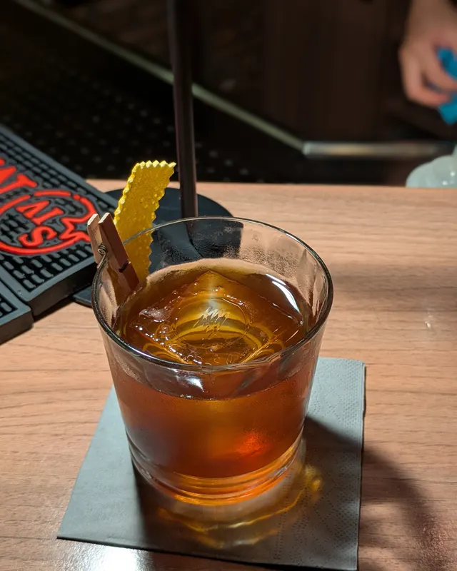 Dark Old Fashioned with a yellow garnish peg on a dark wooden counter